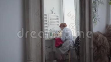 漂亮的红发女人坐在灯房靠窗的桌子边缝衣服。女裁缝在家工作